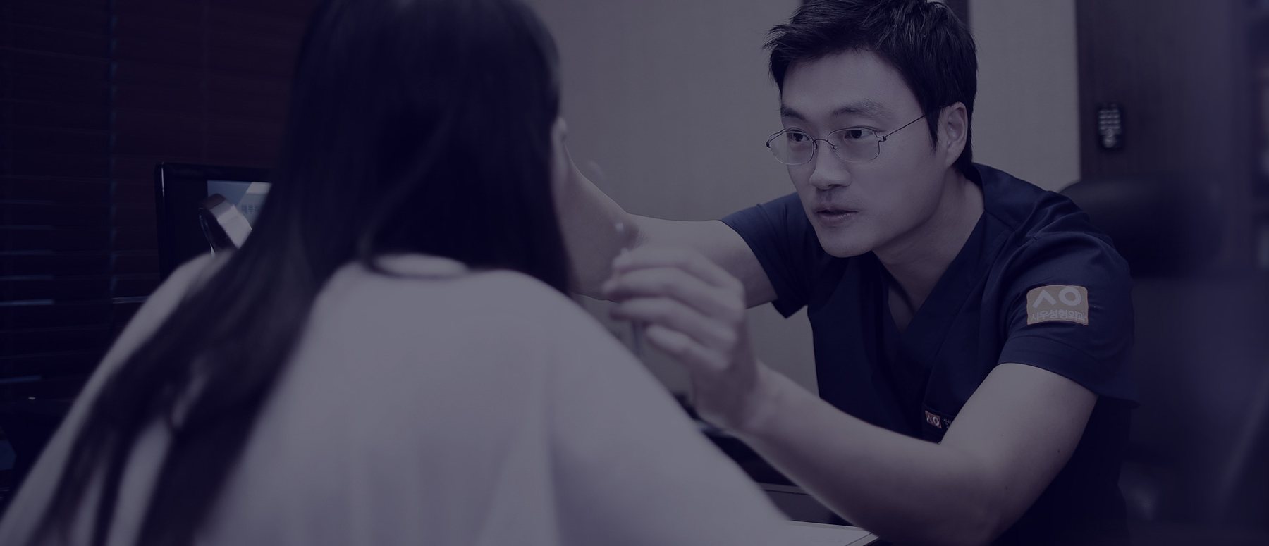 진료실에서 여성 환자(뒷모습)의 얼굴을 손으로 살피며 상담하는 이시우 원장(안경, 남색 수술복)